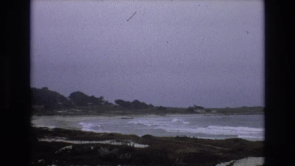 1973 8mm frame of the California coast with white surf across the middle distance, dark rocks in front, and a low wooded shoreline under gray cloud cover.