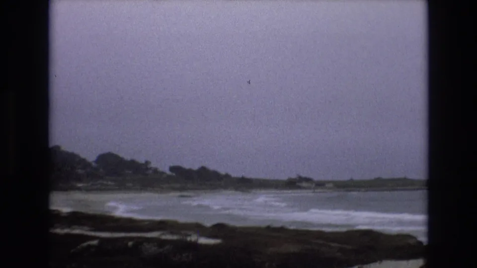 1973 8mm home-movie frame of waves approaching a dark California coastal shoreline, with distant trees and a gray sky across the water.