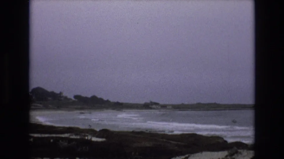 Vintage 8mm view of white surf breaking along the central California coast, with a low distant shoreline and trees under a muted gray sky.