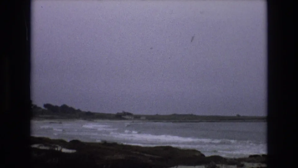 1973 home-movie frame showing foamy waves near a dark shore on the California coast, with faint low buildings and trees across the water.