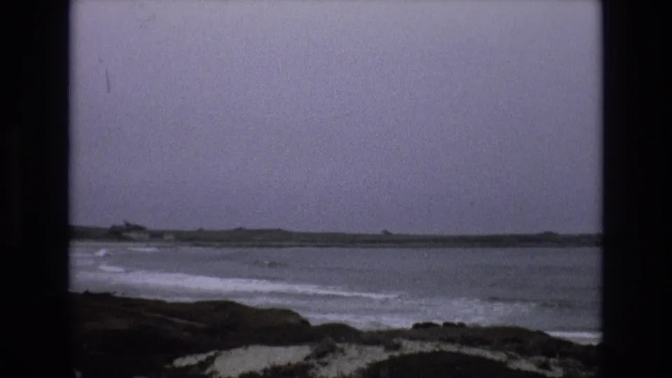 1973 8mm coastal footage shows gray ocean waves breaking near a rocky California shoreline under an overcast sky.