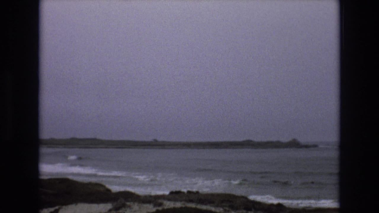 Muted 8mm frame of rough Central Coast surf, a rocky foreground, and a low distant point beneath a gray sky.
