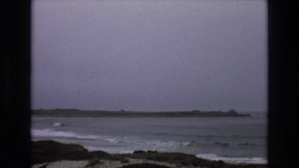 Muted 8mm frame of rough Central Coast surf, a rocky foreground, and a low distant point beneath a gray sky.