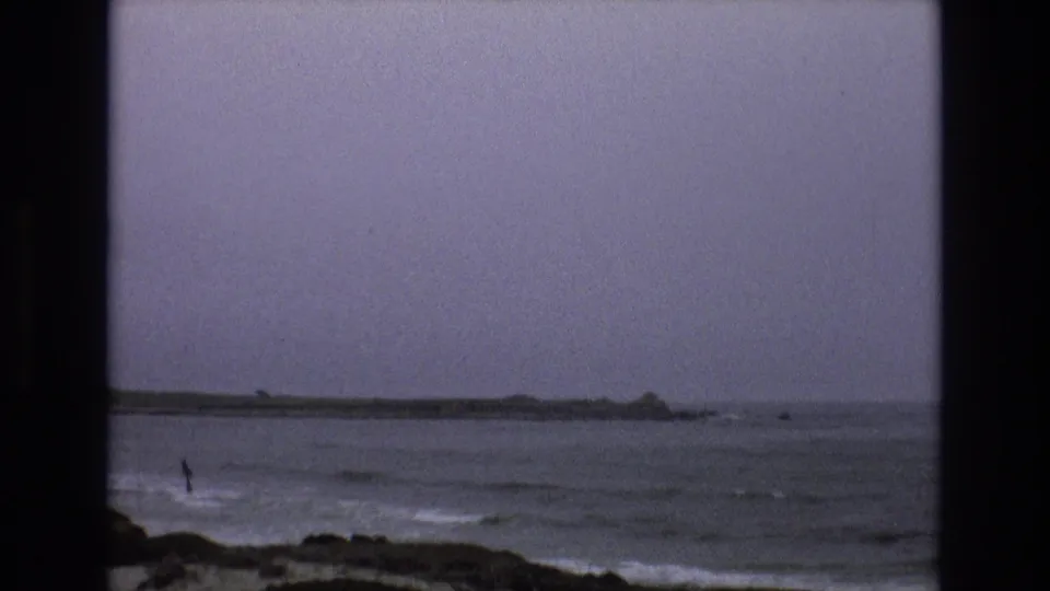 1973 California coastal home-movie frame with choppy gray water, dark rocks, and a low horizon under stormy light.