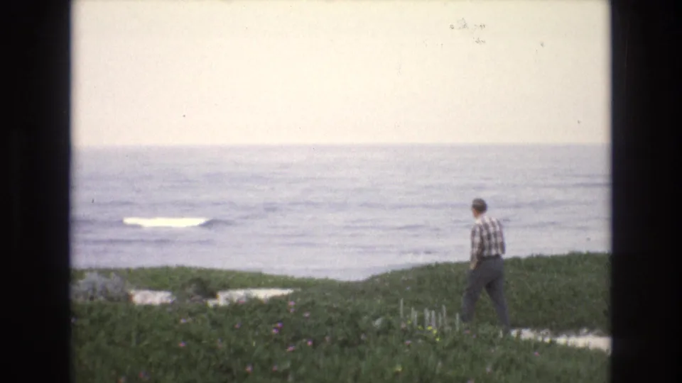 A distant adult figure in a plaid shirt stands on a grassy California coastal bluff, looking toward pale surf and open water in 1973 8mm footage.