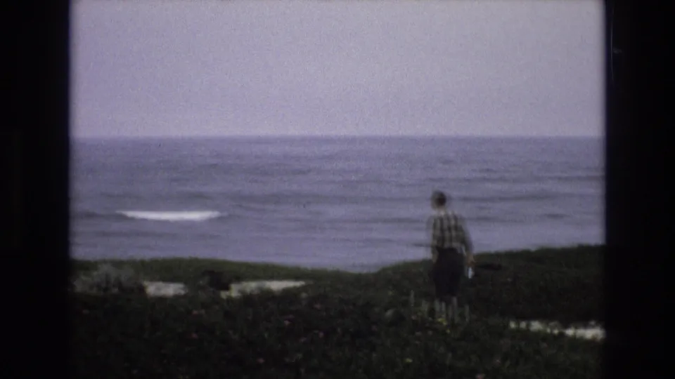 In 1973 California coast home-movie footage, a man in a plaid shirt is seen from behind on a grassy bluff overlooking a restless sea under a pale sky.