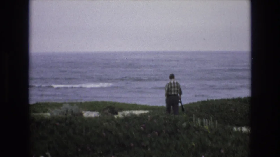 The man appears centered on a grassy Central Coast overlook, filmed from behind against a wide ocean horizon and subdued 8mm color.