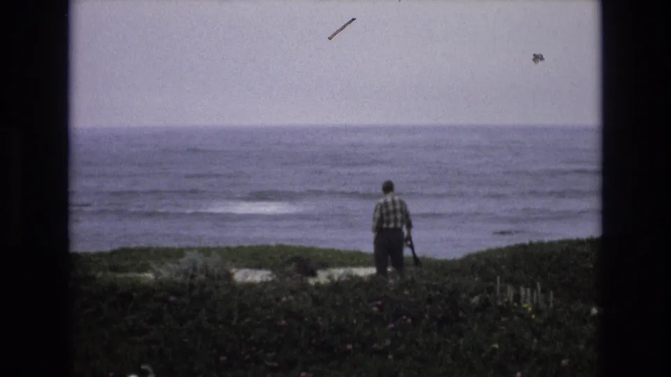 A lone man in plaid stands on low coastal vegetation facing the California shoreline, with subdued waves and visible film specks in the scanned 8mm frame.