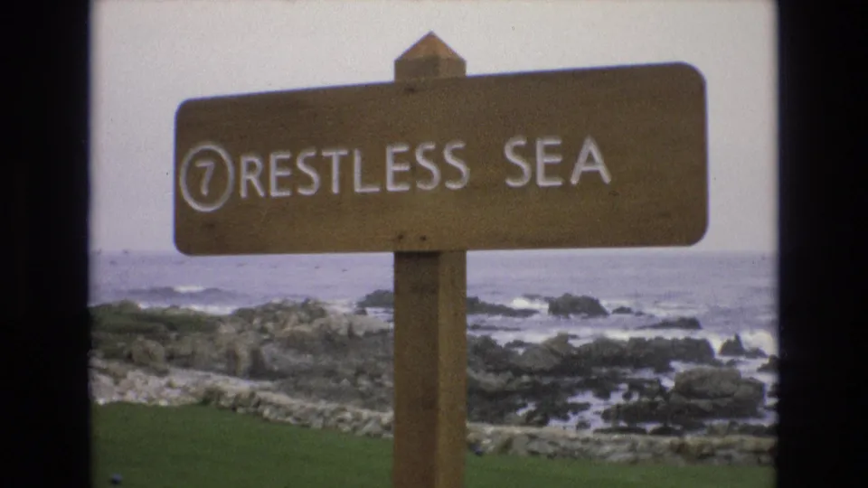 1973 8mm home-movie frame of a wooden "7 RESTLESS SEA" sign on the California coast with rocks, surf, grass, and pale sky behind it.