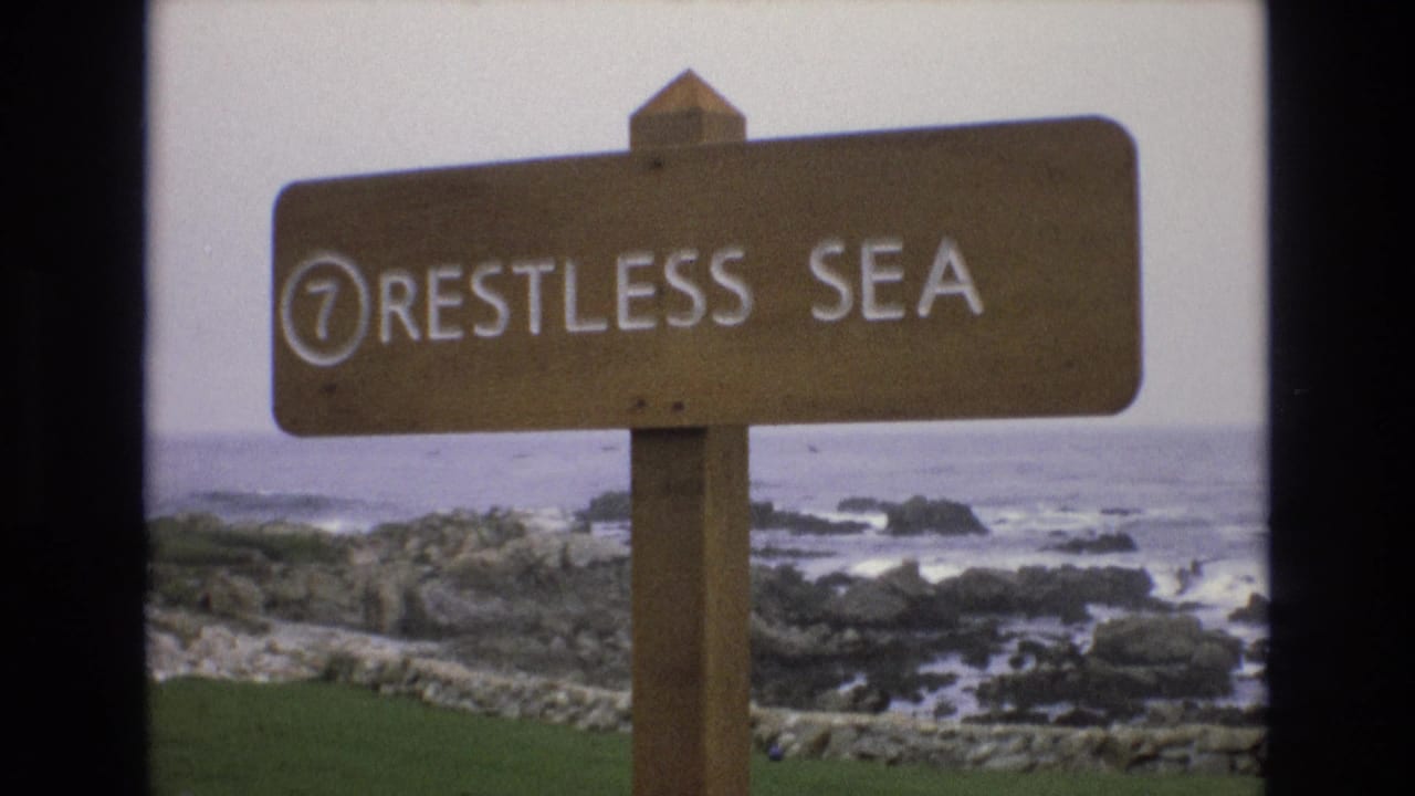 A steady 1973 coastal home-movie view shows the "7 RESTLESS SEA" wooden sign with rocky surf and muted ocean horizon in the background.