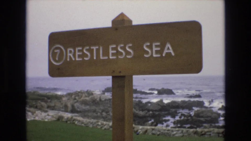 A steady 1973 coastal home-movie view shows the "7 RESTLESS SEA" wooden sign with rocky surf and muted ocean horizon in the background.
