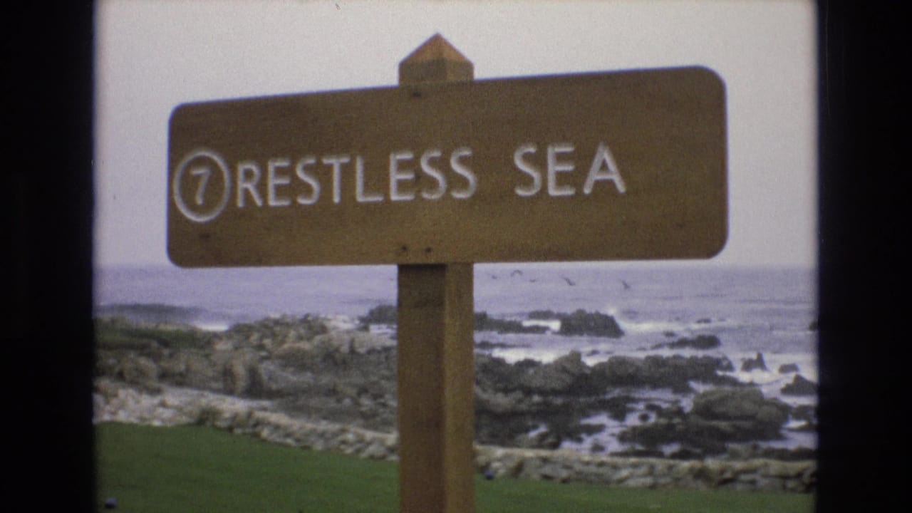 Close view of the Restless Sea sign with shoreline rocks and water behind it.