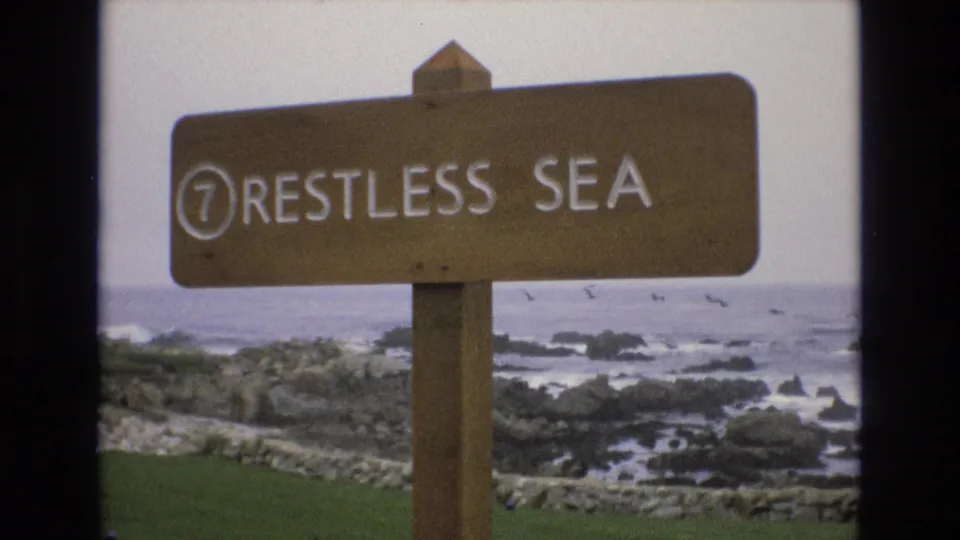 Final 1973 8mm frame of the wooden Restless Sea sign on the California coast, with rocky surf, several distant birds, and pale daylight in the background.