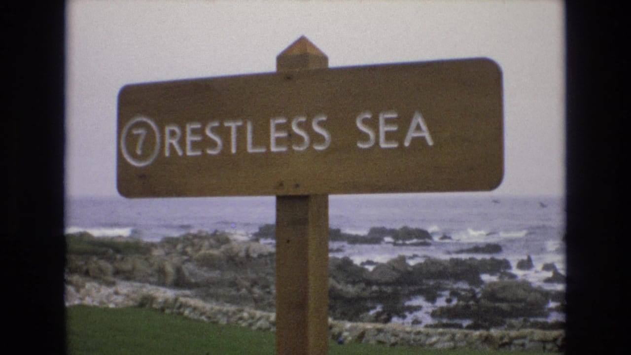A second 1973 8mm view holds on the wooden 7 RESTLESS SEA sign, with pale ocean water, dark rocks, and grey sky in the background.