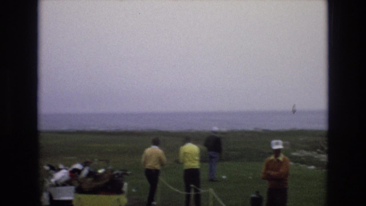 Golfers and spectators stand on a coastal green under a hazy sky.