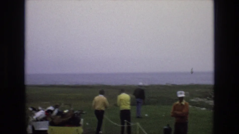 1973 8mm frame showing several men on or near a coastal golf green, with a rope boundary, golf bags, ocean horizon, and foggy Central Coast California weather.