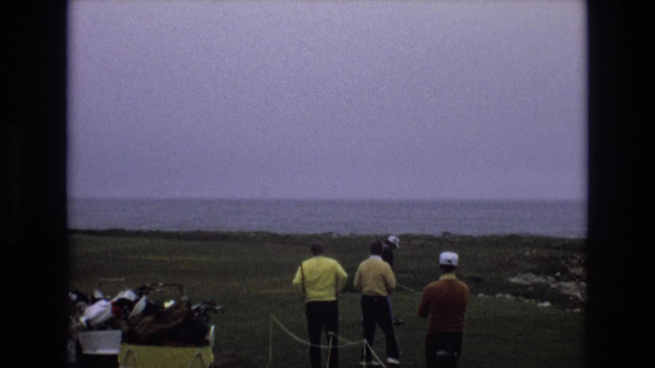 1973 Central Coast California 8mm frame showing adult golfers in bright sweaters near a yellow golf cart on a grassy tee area overlooking distant ocean water.