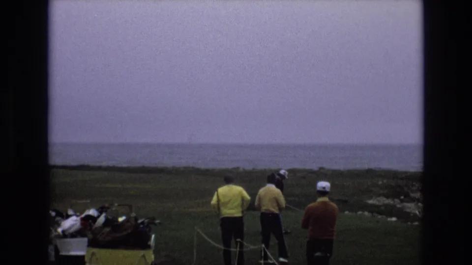 1973 Central Coast California 8mm frame showing adult golfers in bright sweaters near a yellow golf cart on a grassy tee area overlooking distant ocean water.