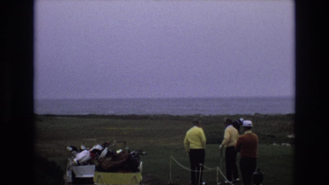 Golfers in yellow and orange sweaters stand beside a loaded golf cart with the coastline behind them.