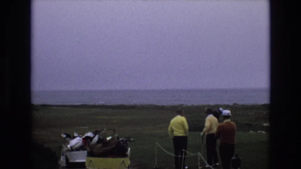 1973 Central Coast California home-movie frame of golfers standing near a club-filled yellow cart on a coastal fairway under a hazy overcast sky.