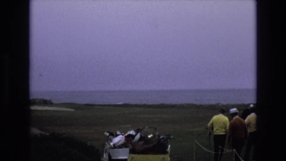 1973 Central Coast California 8mm frame showing adult golfers moving from a yellow cart toward the fairway, with low coastal land and the ocean in the background.