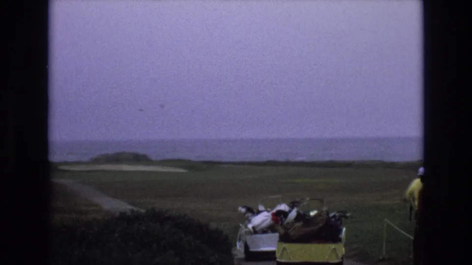 1973 Central Coast California home-movie frame with a yellow golf cart and bags at the edge of a coastal course, while golfers continue out of frame beside an ocean-view fairway.