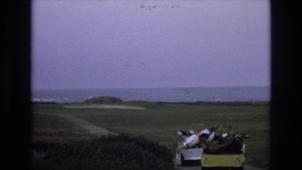 Coastal golf fairway with sand traps, ocean horizon, and a golf cart with clubs in the foreground.