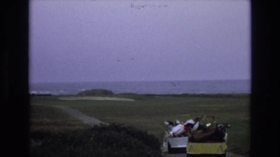 1973 8mm home-movie frame showing a California coastal golf course with fairway, sand traps, ocean horizon, and a golf cart loaded with clubs near the lower edge.