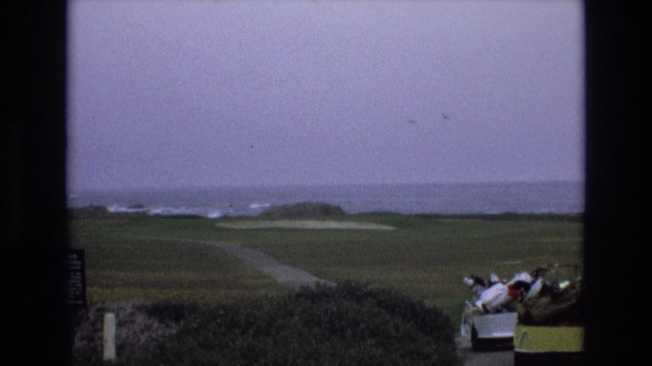 1973 California coastal golf-course view with a winding path across the fairway, pale sand traps, surf in the distance, and a golf cart partly visible at right.