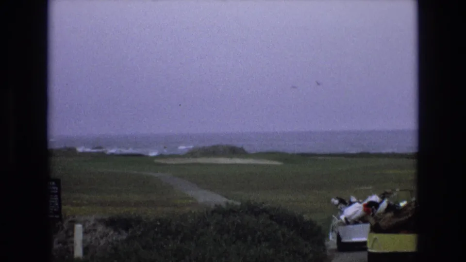 1973 California coastal golf-course view with a winding path across the fairway, pale sand traps, surf in the distance, and a golf cart partly visible at right.
