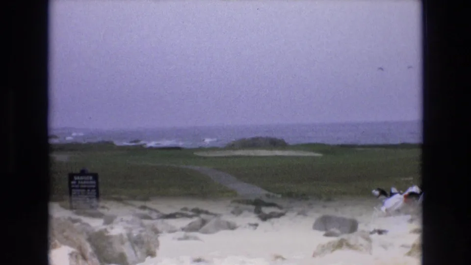 1973 8mm frame of a California coastal fairway with a small course sign, sand areas, ocean surf beyond the green, and part of a golf cart entering the lower right.