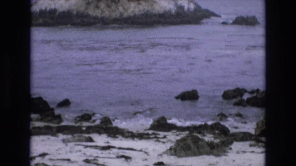 1973 California coast home-movie frame showing rocky shoreline, shallow ocean water, scattered rocks, and surf beyond the golf-course setting.