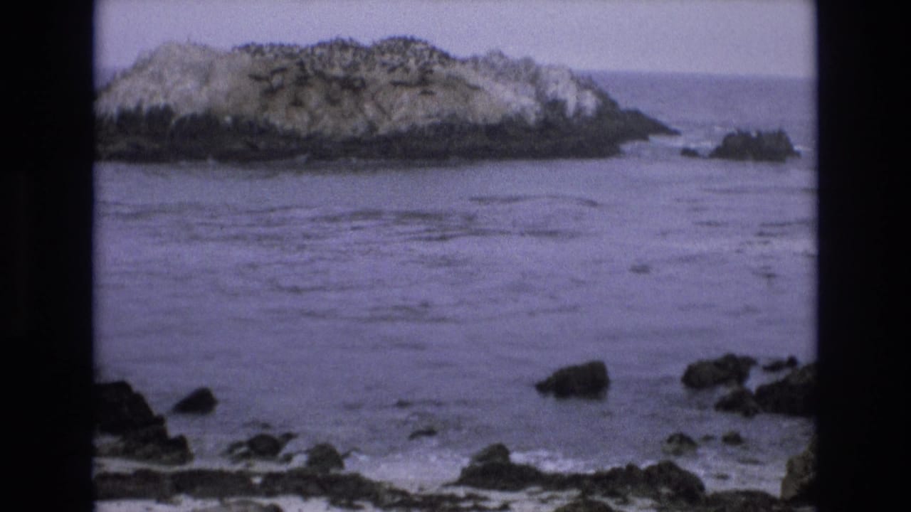 A 1973 8mm home-movie frame of the California coast shows a rocky offshore sea stack across gray-blue water, with dark shoreline rocks in the foreground.
