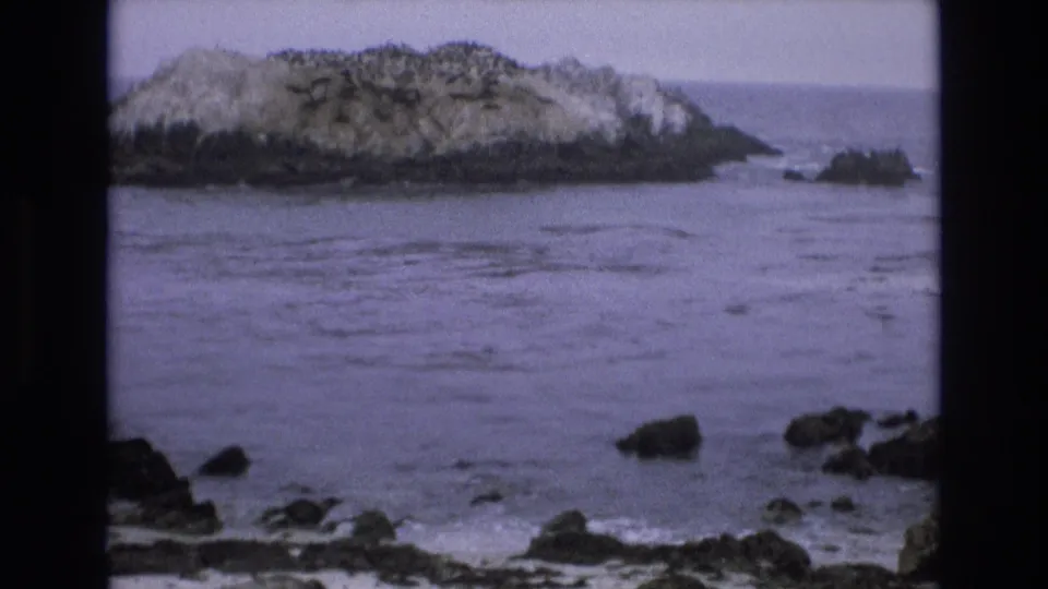 A 1973 8mm home-movie frame of the California coast shows a rocky offshore sea stack across gray-blue water, with dark shoreline rocks in the foreground.