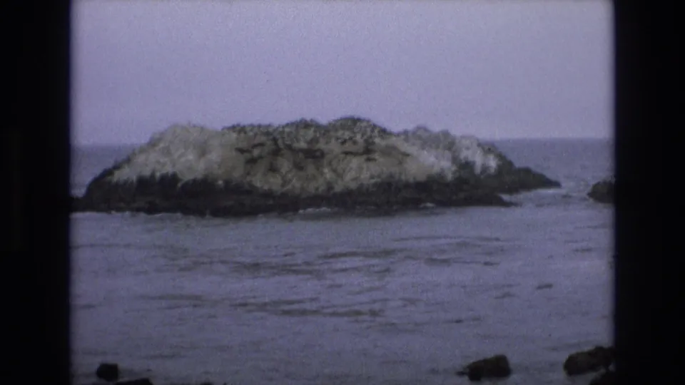 A closer 1973 coastal frame shows the offshore rock filling more of the view, with subdued ocean ripples and sparse shoreline stones along the bottom edge.