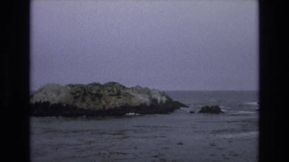 1973 8mm Central Coast California frame showing a low rocky offshore islet with calm water, small waves, and dark film borders.