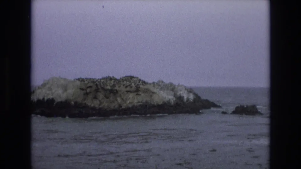 1973 8mm Central Coast California view of a rugged islet with a smaller rock outcrop nearby and soft waves around the base.