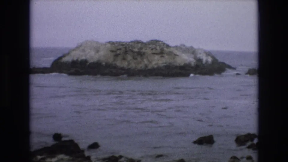 1973 8mm Central Coast California frame showing a low rocky island offshore with calm water and shoreline rocks in the foreground.