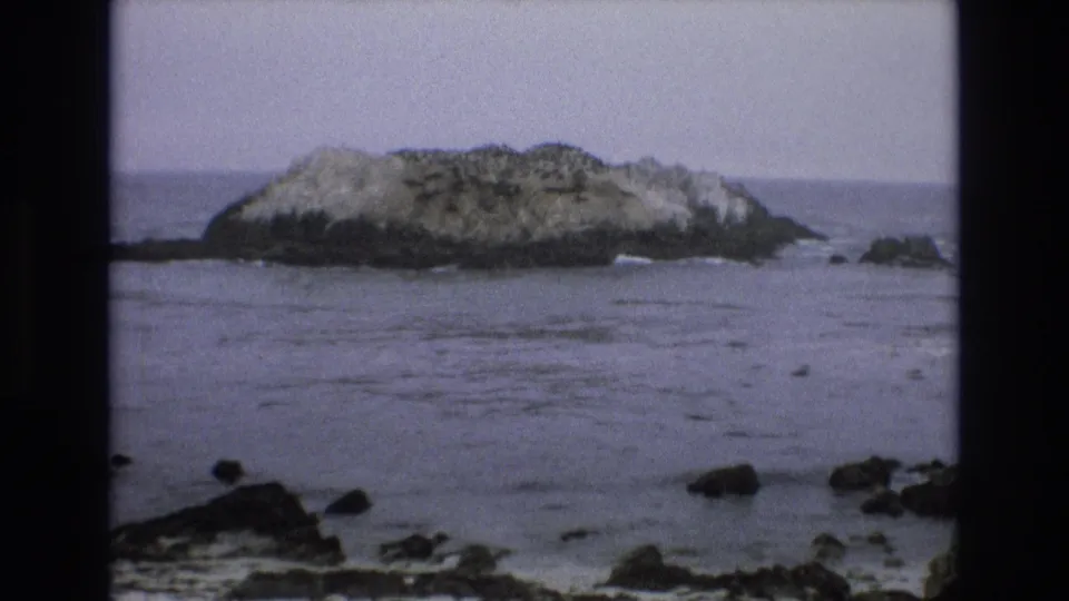 1973 8mm coastal view of a small rock islet in the ocean, with scattered foreground rocks and a muted horizon line.