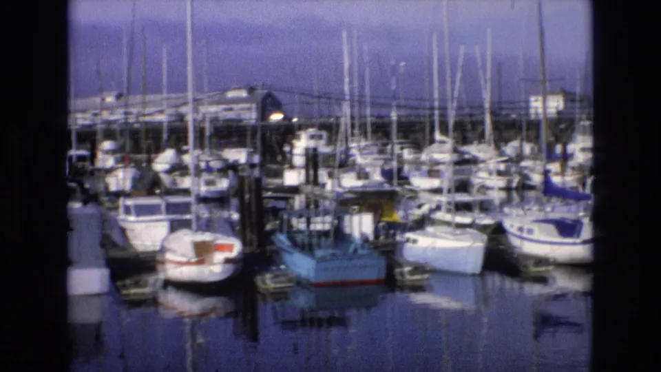 1973 California coastal home-movie frame showing a marina with moored sailboats and small boats, vertical masts, dock structures, reflections, and a purple-tinged sky.