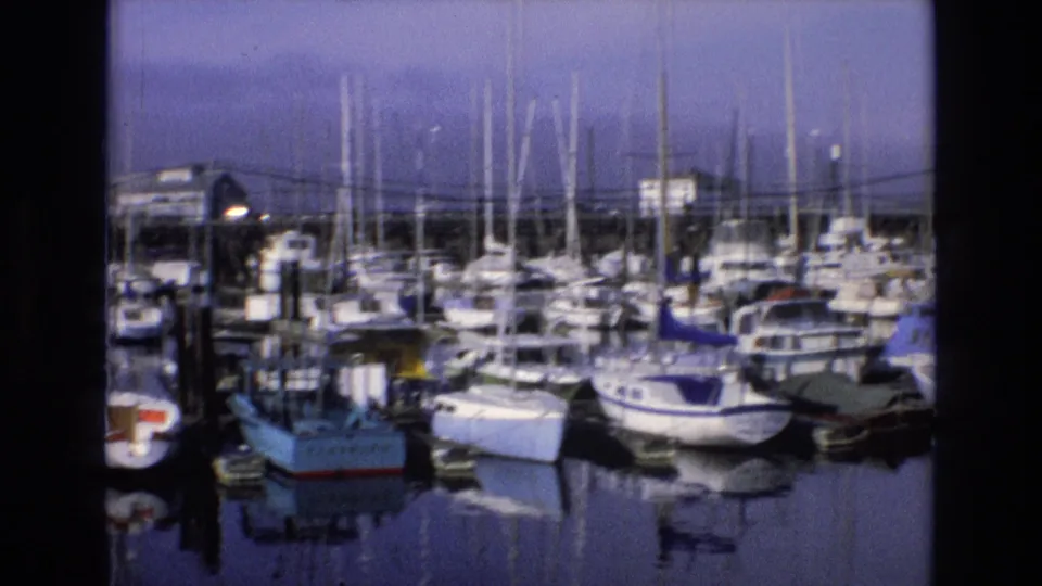 1973 Central Coast California home-movie frame showing a crowded marina with sailboat masts, cabin boats, dock edges, and calm reflective water.