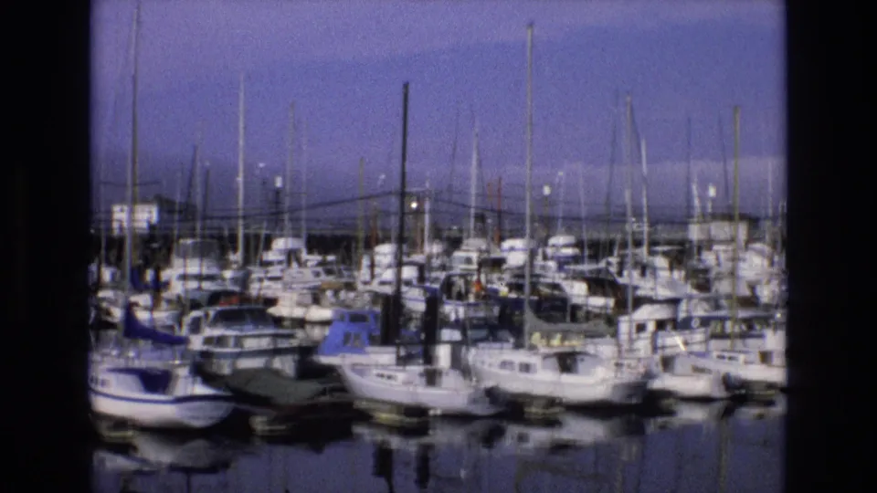Home-movie frame from the 1973 Central Coast California clip showing moored boats, docked cabins, vertical rigging, and still water reflections.