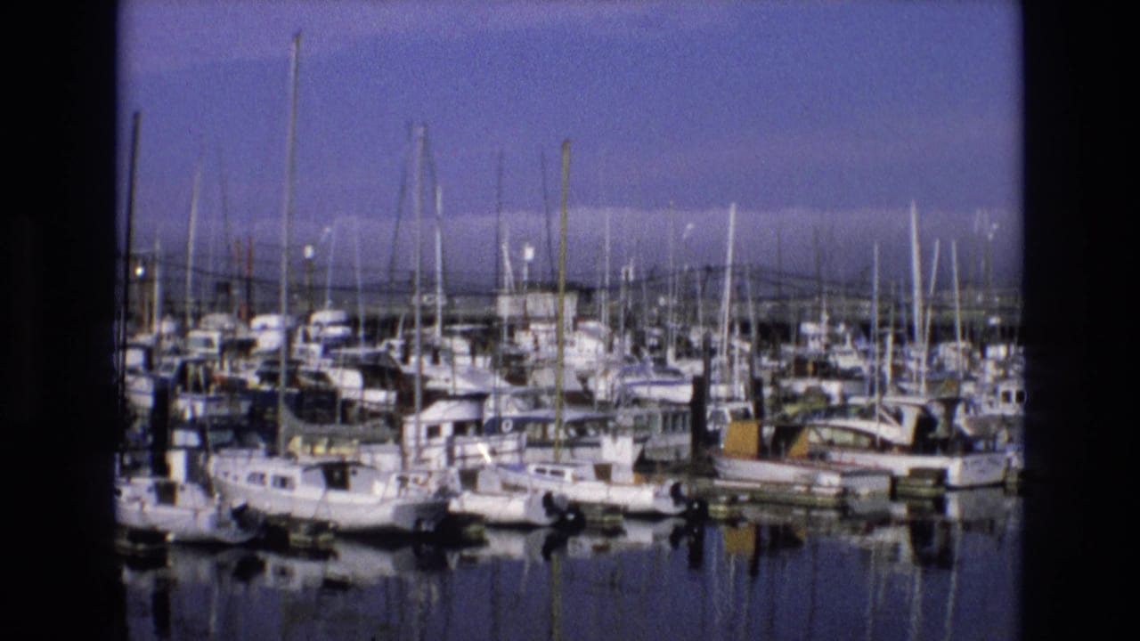 1973 8mm view of a Central Coast California marina with closely docked boats, tall masts, calm reflective water, and dark film-frame borders.