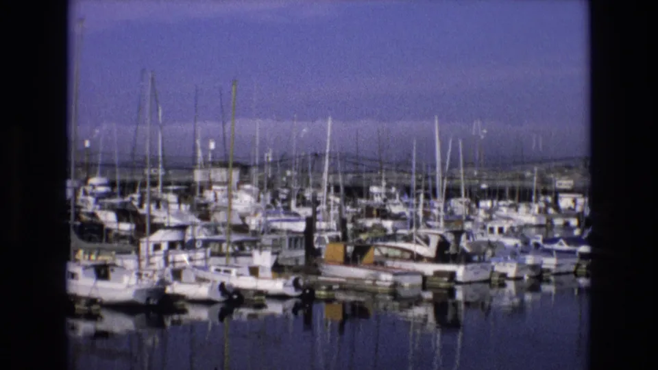 1973 Central Coast California marina scene showing rows of docked small craft, sailboat masts, still water reflections, and a soft 8mm home-movie look.