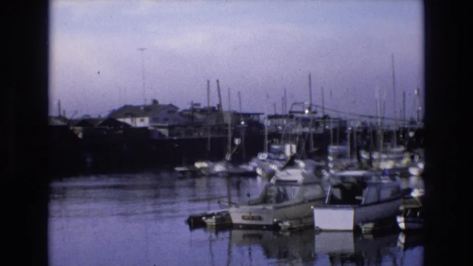 1973 8mm frame of a California coastal marina with small boats in the foreground, waterfront buildings beyond, many masts, and calm harbor water.