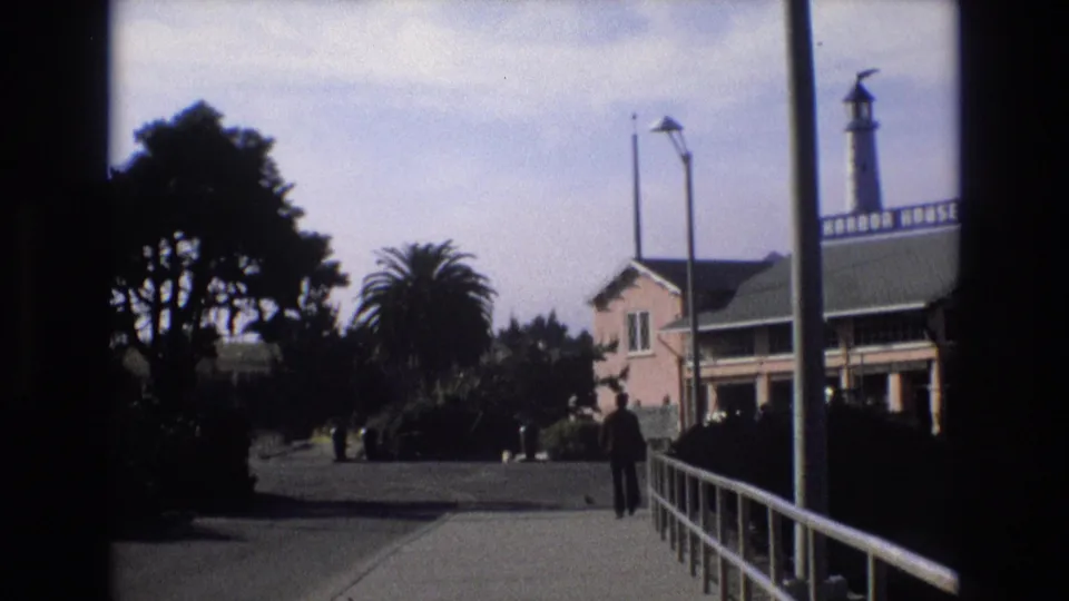 1973 California waterfront walkway where a man appears to walk a dog near trees, railings, and a Harbor House building with a lighthouse-style tower.