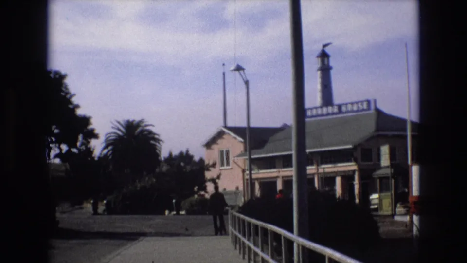 1973 Central Coast California home-movie frame of a waterfront path with a man and dog, railings, trees, and Harbor House signage on a harbor-side building.
