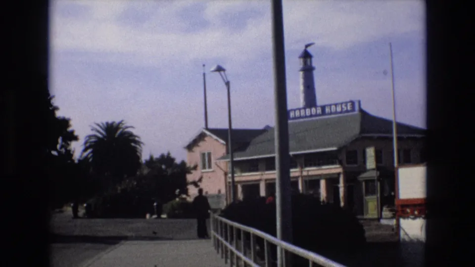1973 8mm frame of a Central Coast California waterfront scene showing the Harbor House building, its lighthouse-style tower, walkway railings, palm trees, and a distant adult figure.