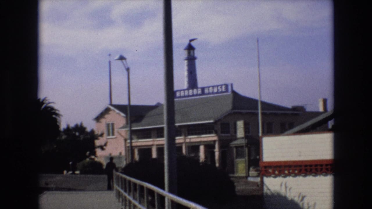 1973 Central Coast California harbor view with the Harbor House sign on a waterfront building, a lighthouse-style tower, palm trees, railing, and a person walking in the distance.
