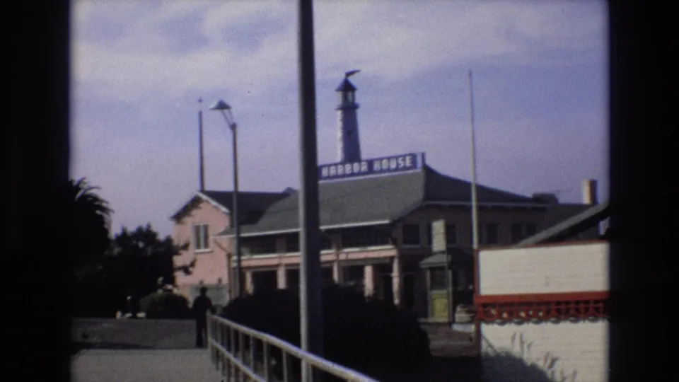 1973 Central Coast California harbor view with the Harbor House sign on a waterfront building, a lighthouse-style tower, palm trees, railing, and a person walking in the distance.
